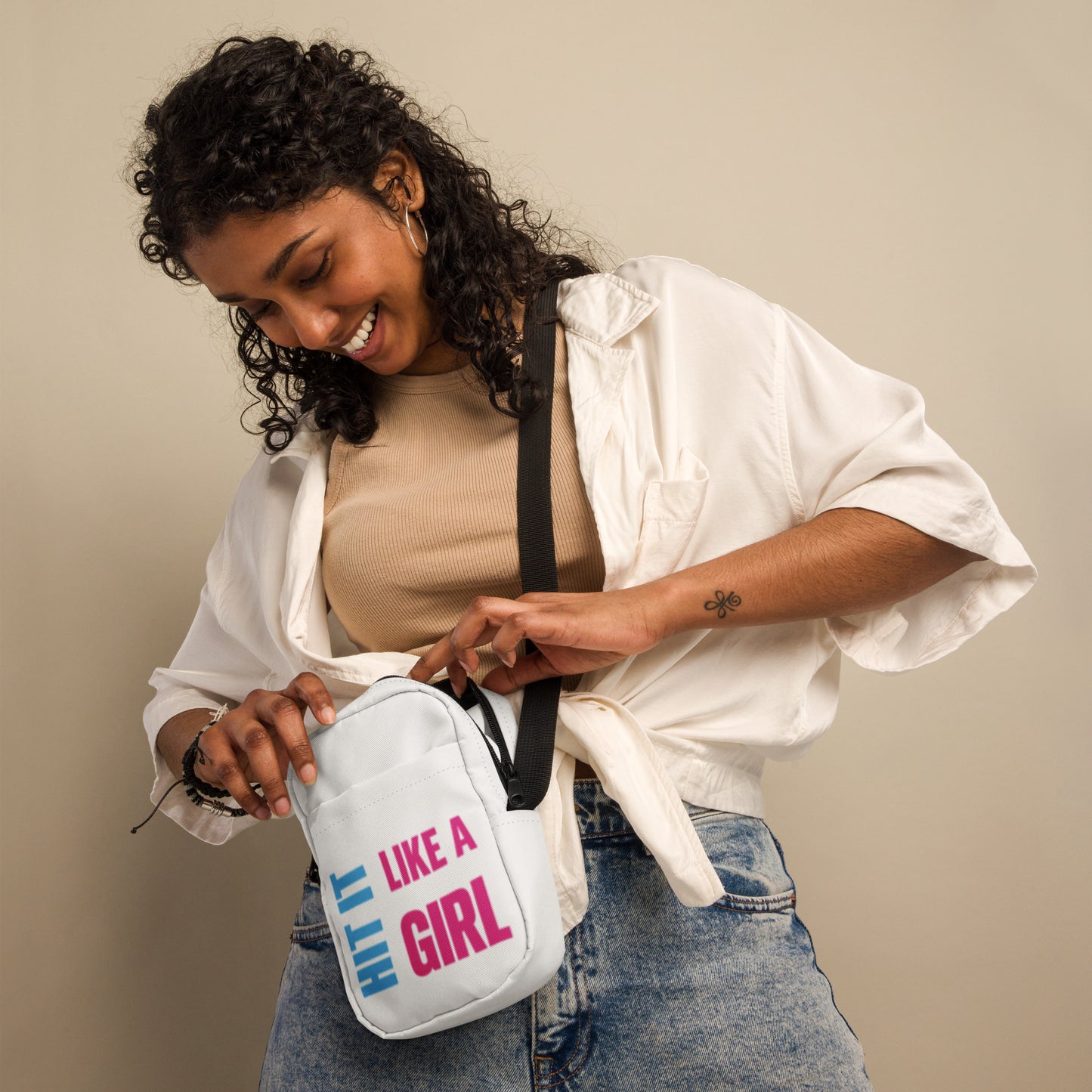 Woman wearing a white Hit It Like a Girl crossbody bag with bold pink and blue logo — compact, empowering gear for sporty women on the move.
