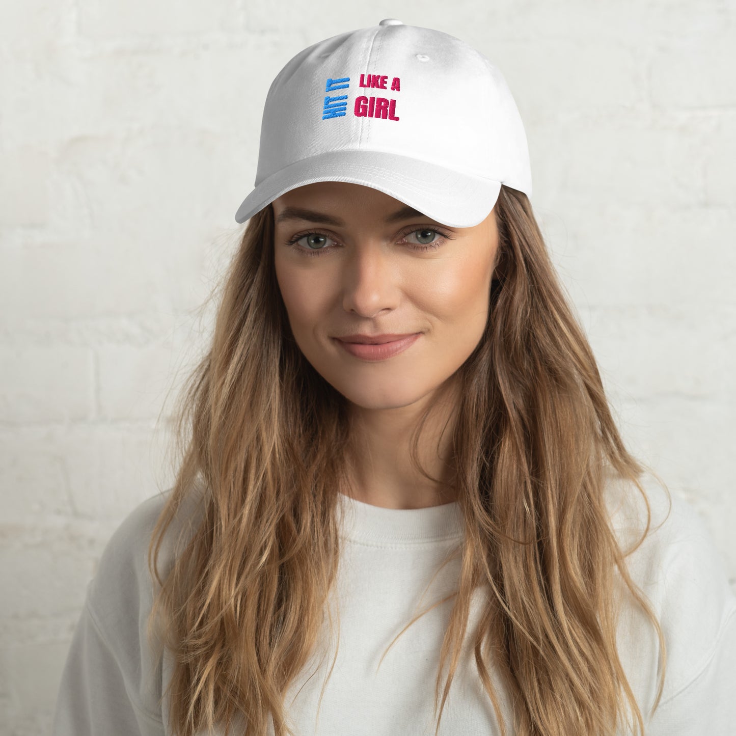 Close-up of a white "Hit It Like a Girl" baseball cap featuring bold blue and pink embroidered text, worn by a woman with long wavy hair and a neutral background.