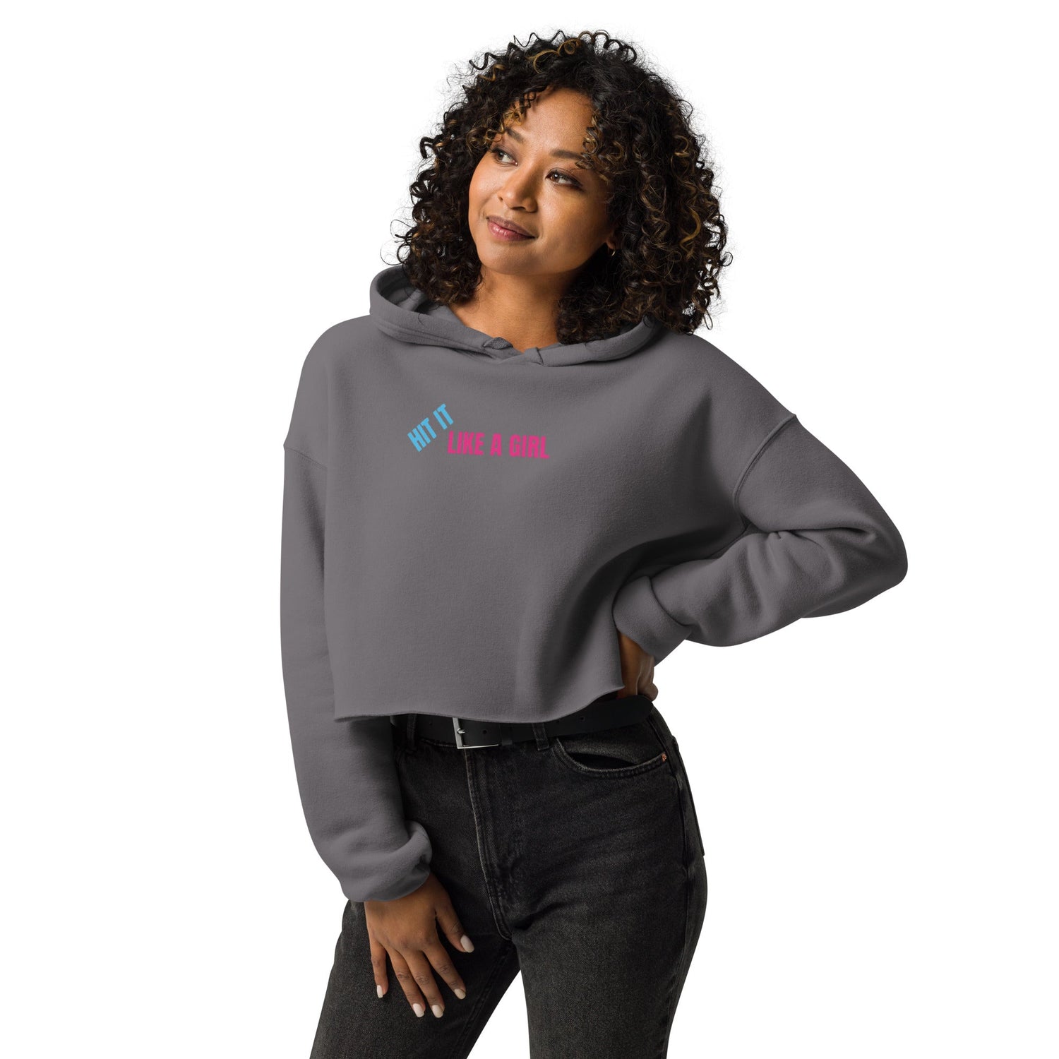 Woman wearing a gray Hit It Like a Girl cropped hoodie with bold pink and blue logo, styled casually with jeans for a sporty, empowering look.