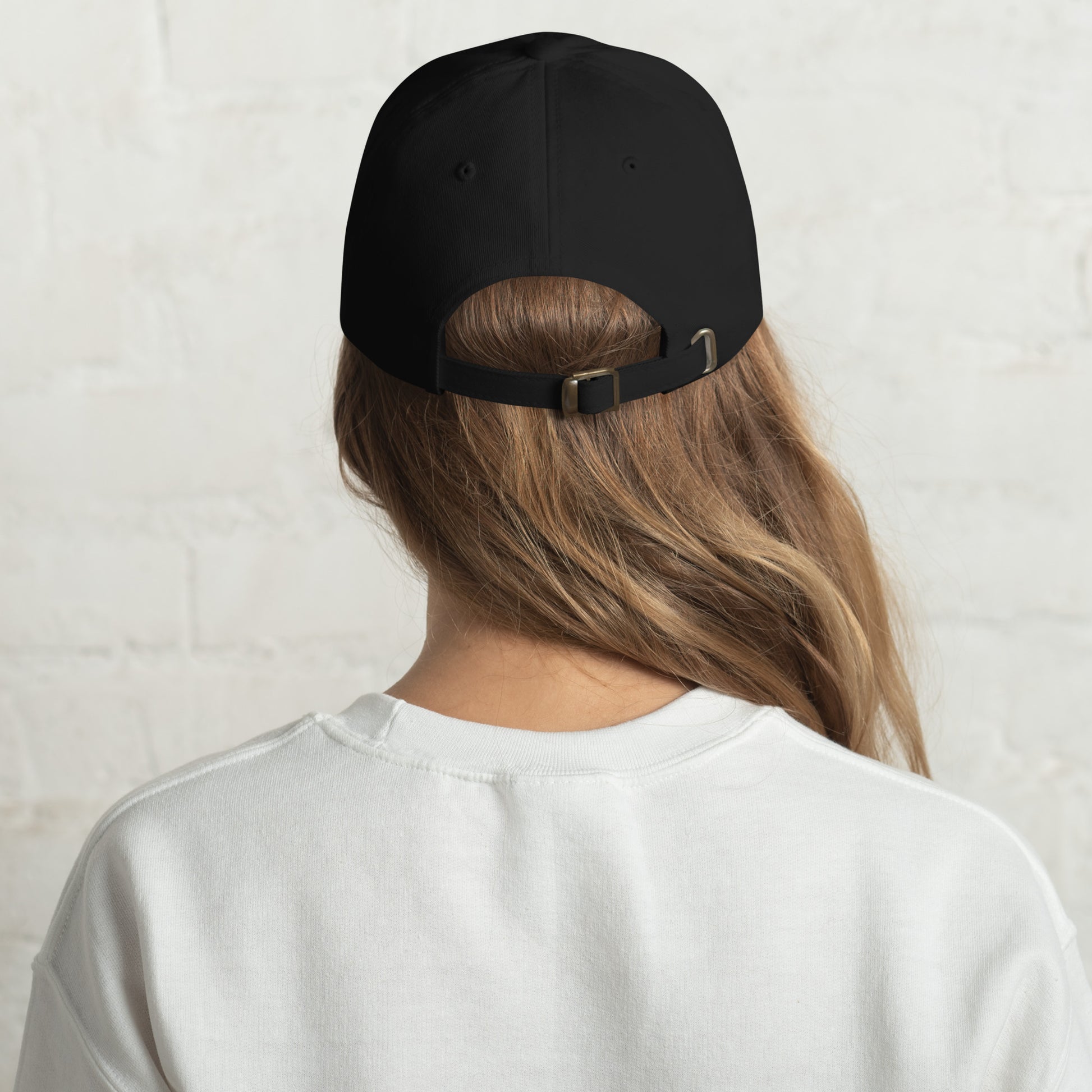 Close-up of a black "Hit It Like a Girl" baseball cap featuring bold blue and pink embroidered text, worn by a woman with long wavy hair and a neutral background.