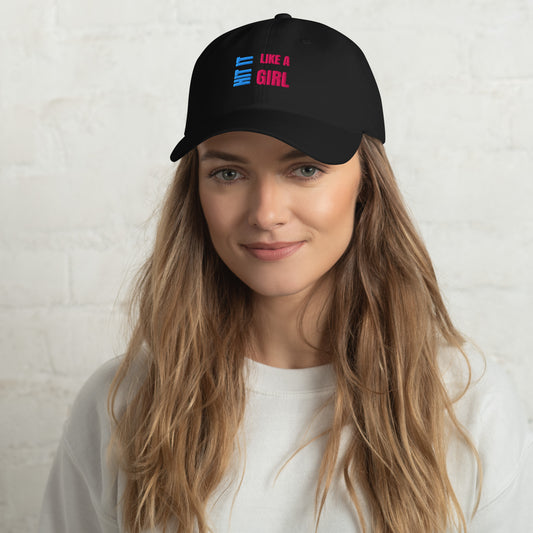Close-up of a black "Hit It Like a Girl" baseball cap featuring bold blue and pink embroidered text, worn by a woman with long wavy hair and a neutral background.