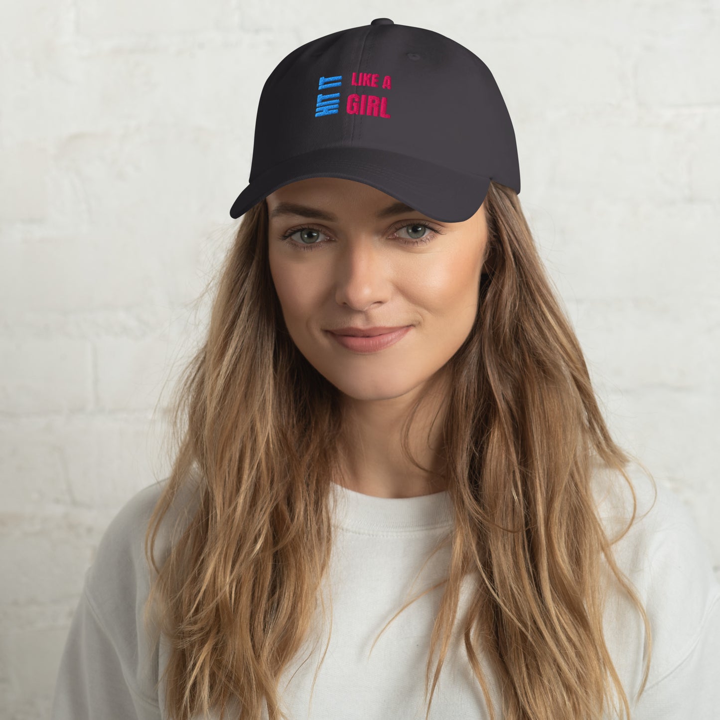 Close-up of a dark gray "Hit It Like a Girl" baseball cap featuring bold blue and pink embroidered text, worn by a woman with long wavy hair and a neutral background.
