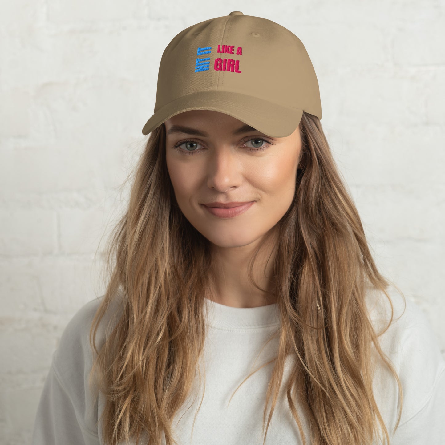 Close-up of a khaki "Hit It Like a Girl" baseball cap featuring bold blue and pink embroidered text, worn by a woman with long wavy hair and a neutral background.