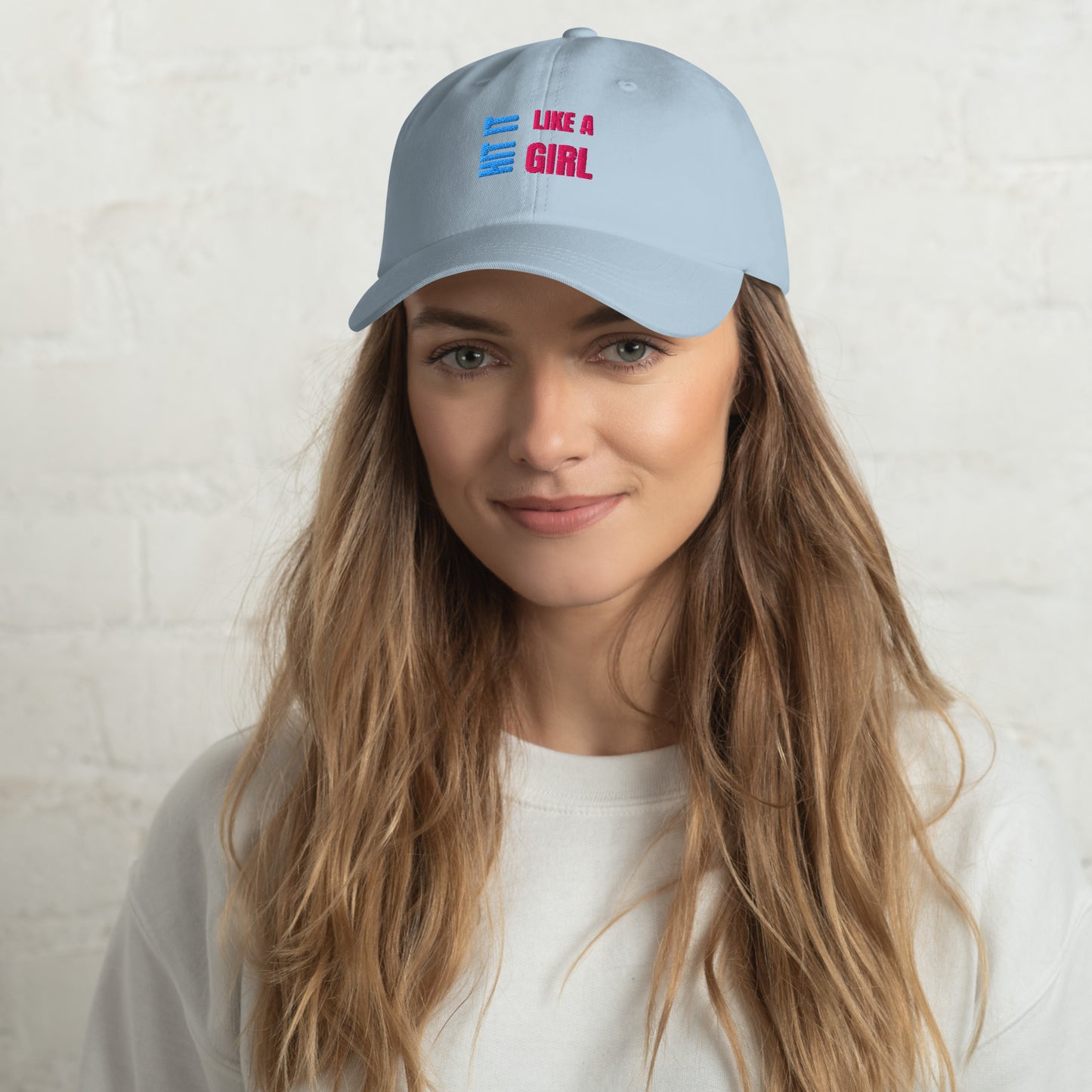 Close-up of a light blue "Hit It Like a Girl" baseball cap featuring bold blue and pink embroidered text, worn by a woman with long wavy hair and a neutral background.