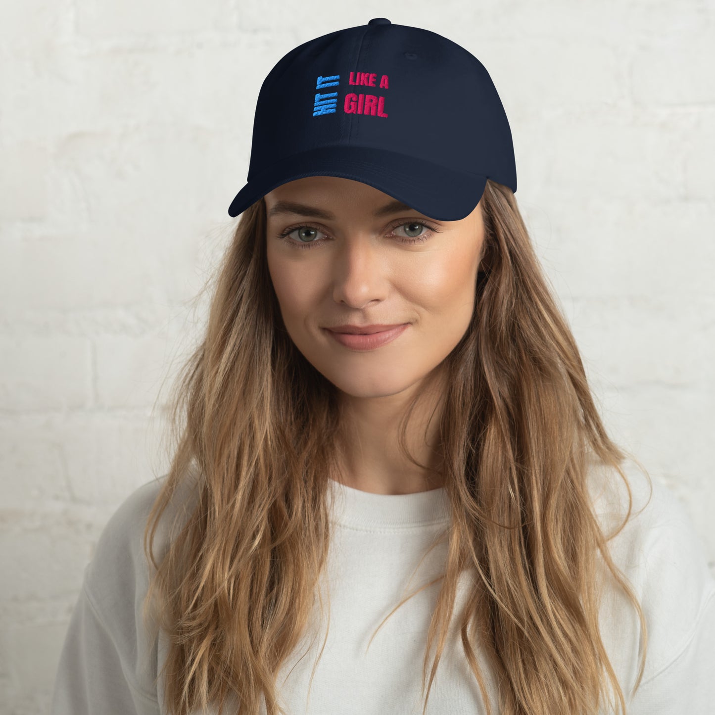 Close-up of a navy "Hit It Like a Girl" baseball cap featuring bold blue and pink embroidered text, worn by a woman with long wavy hair and a neutral background.