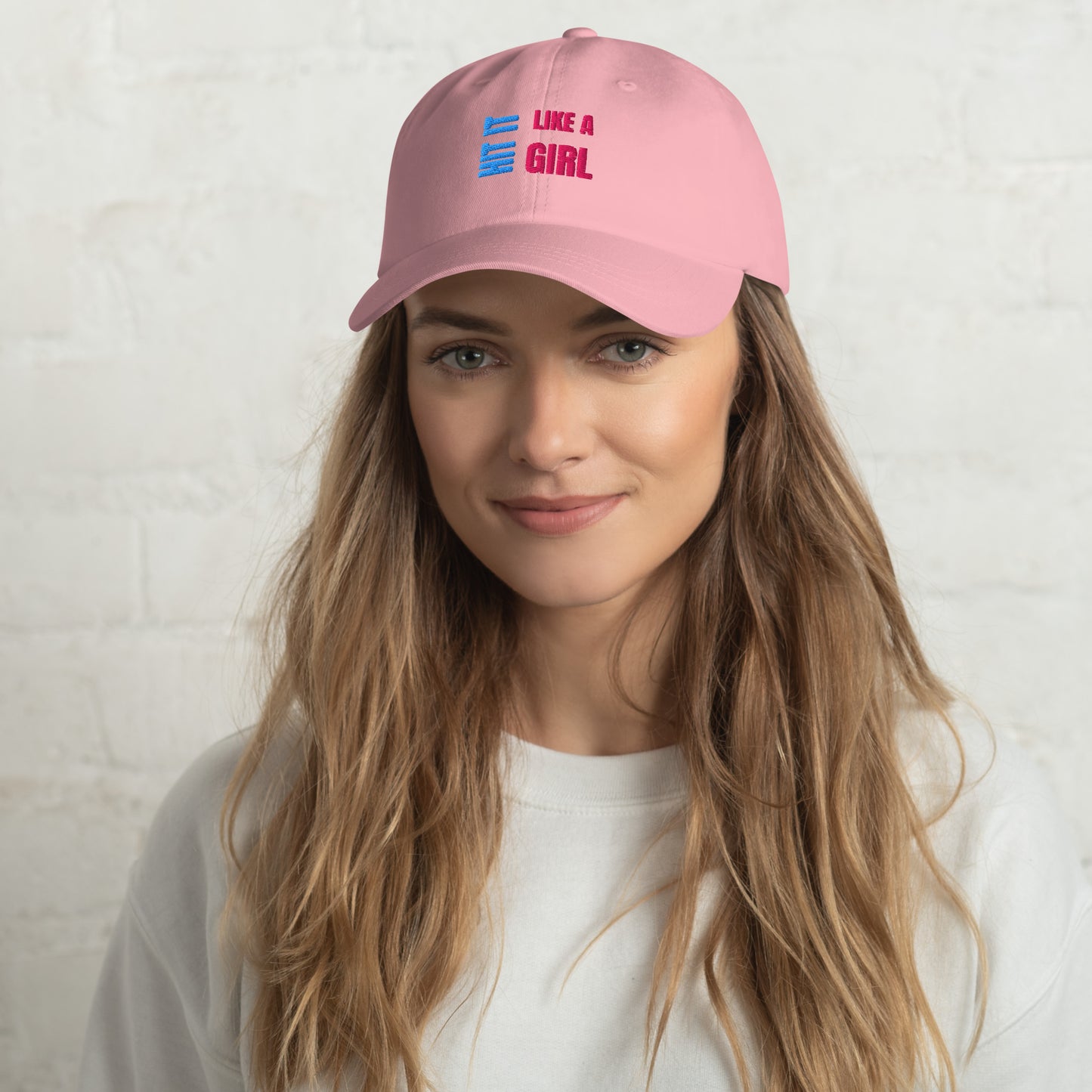 Close-up of a pink "Hit It Like a Girl" baseball cap featuring bold blue and pink embroidered text, worn by a woman with long wavy hair and a neutral background.