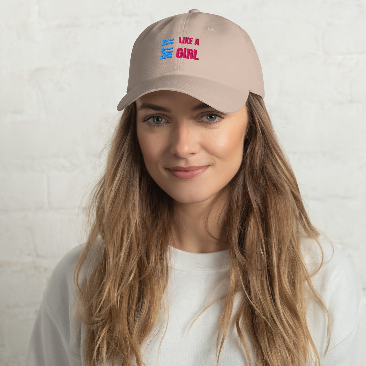 Close-up of a stone "Hit It Like a Girl" baseball cap featuring bold blue and pink embroidered text, worn by a woman with long wavy hair and a neutral background.