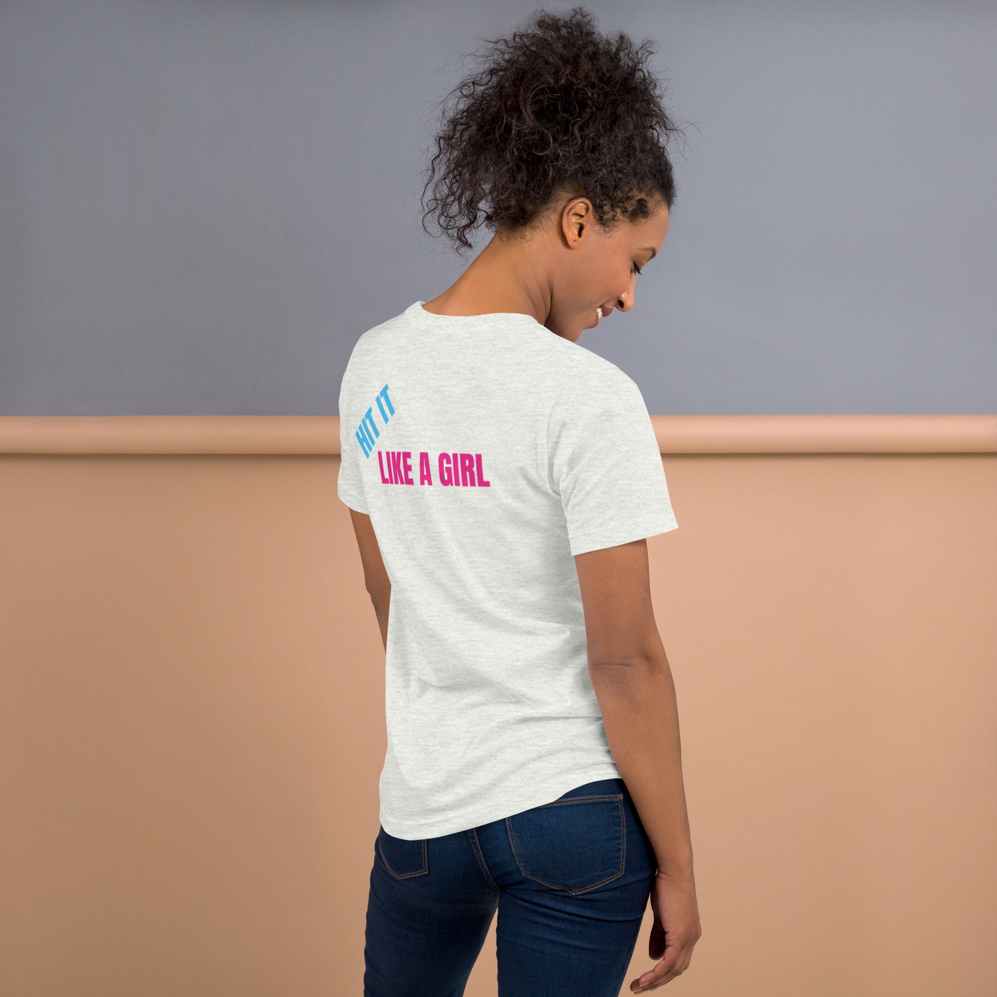 Woman wearing an ash "Hit It Like a Girl" athletic tee with bold blue and pink lettering on the back, standing confidently in casual jeans.
