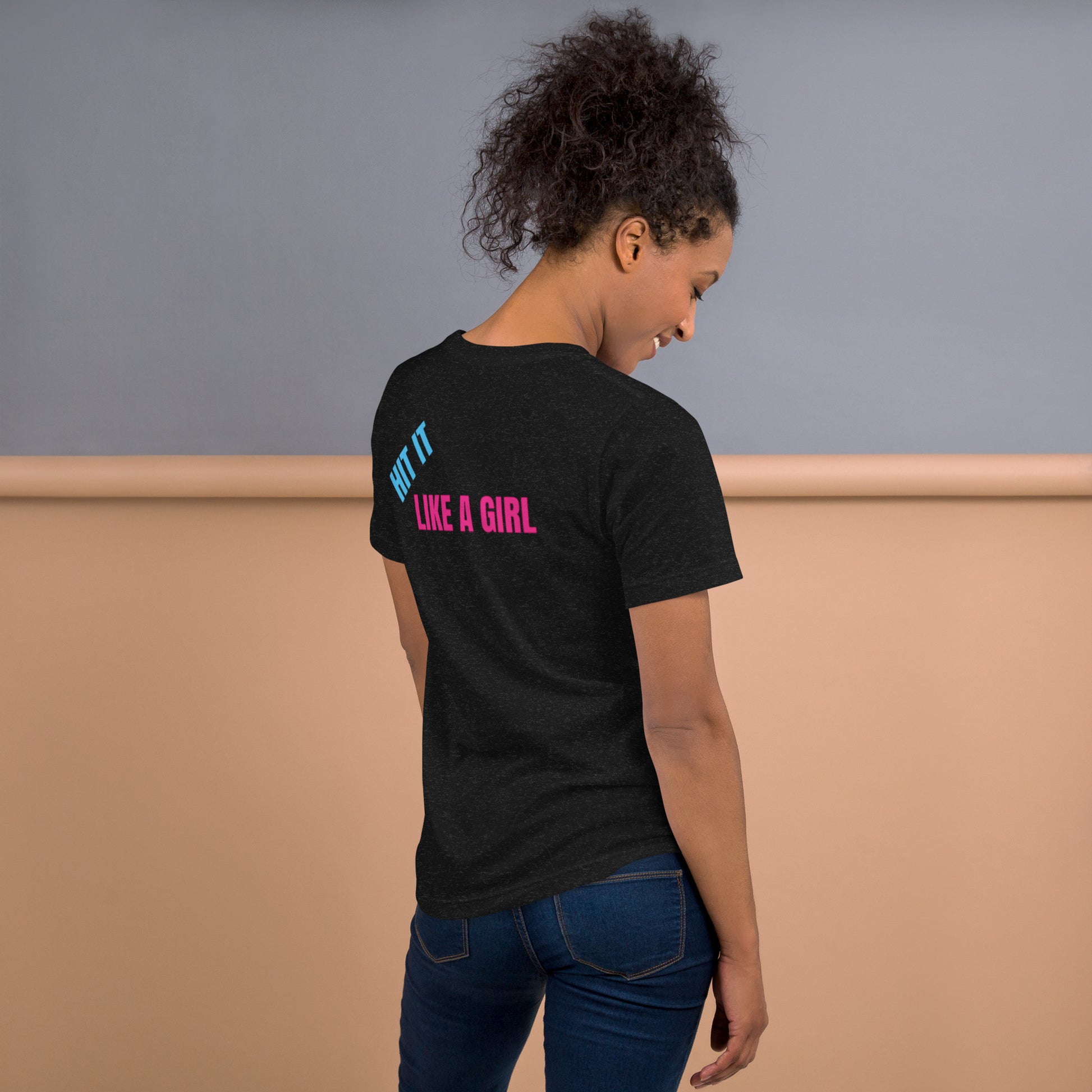 Woman wearing a black heather "Hit It Like a Girl" athletic tee with bold blue and pink lettering on the back, standing confidently in casual jeans.