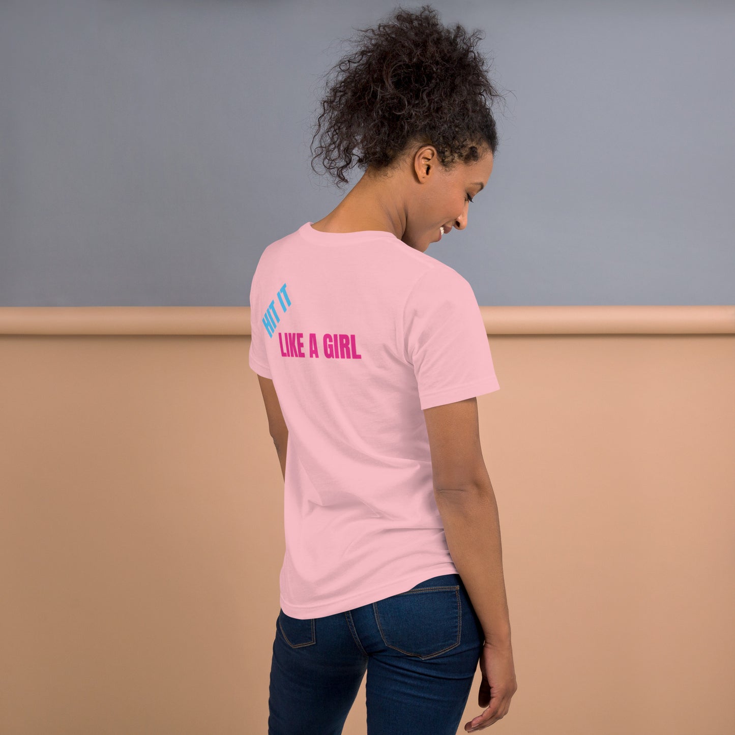 Woman wearing a pink "Hit It Like a Girl" athletic tee with bold blue and pink lettering on the back, standing confidently in casual jeans.