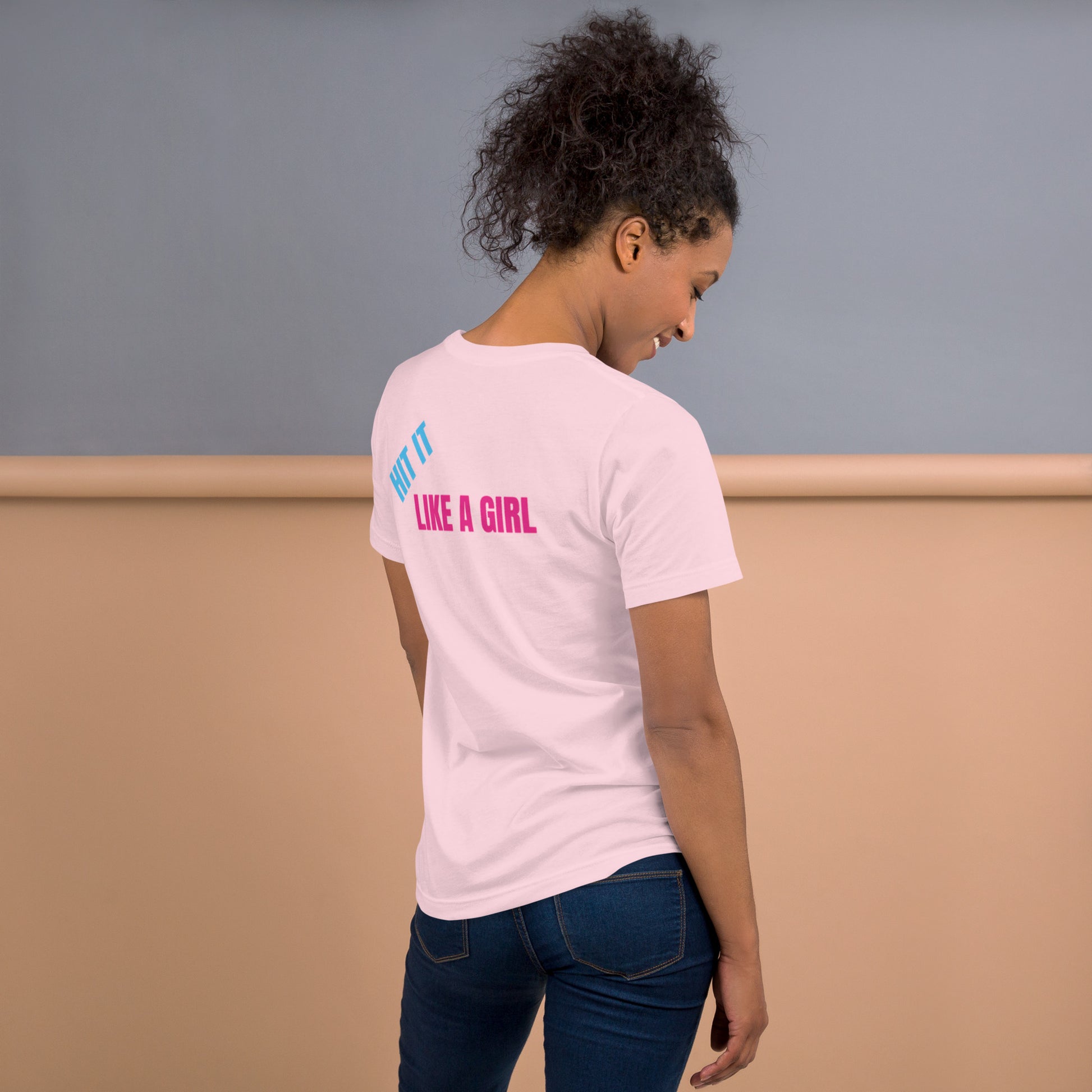 Woman wearing a soft pink "Hit It Like a Girl" athletic tee with bold blue and pink lettering on the back, standing confidently in casual jeans.