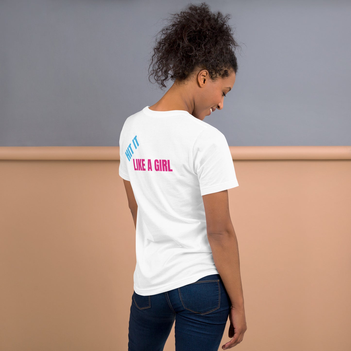 Woman wearing a white "Hit It Like a Girl" athletic tee with bold blue and pink lettering on the back, standing confidently in casual jeans.